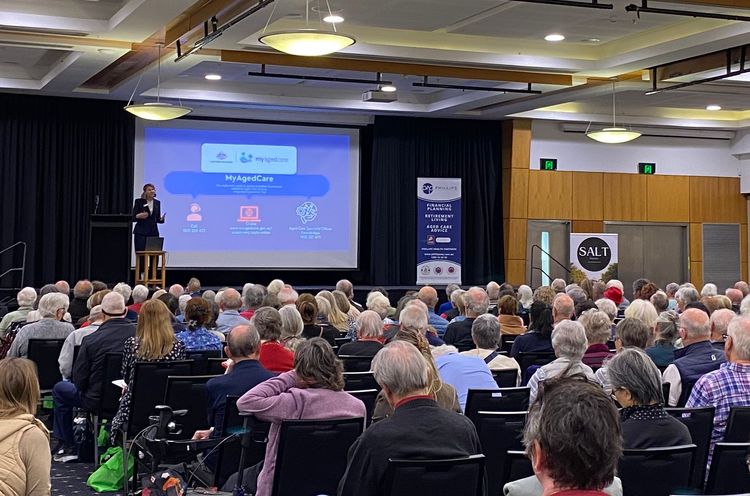 Man presenting to a large audience at a conference, with a screen displaying a logo.
