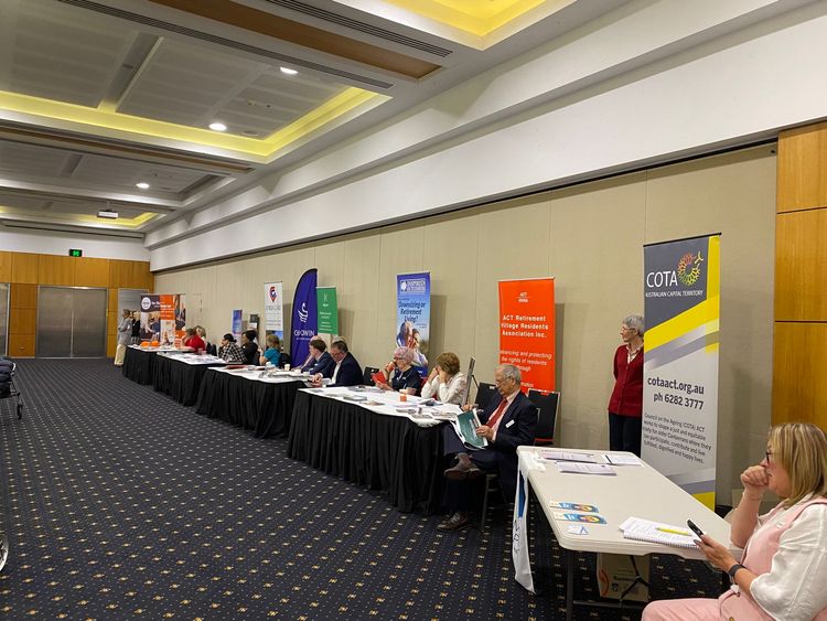 Conference room with presenters behind tables, banners, and audience.