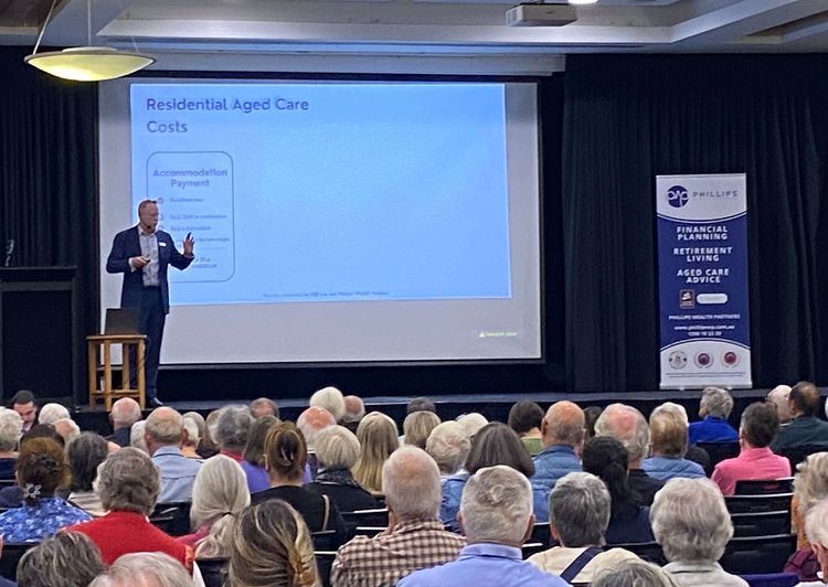 Man presenting to an audience about aged care costs. The setting is an auditorium.