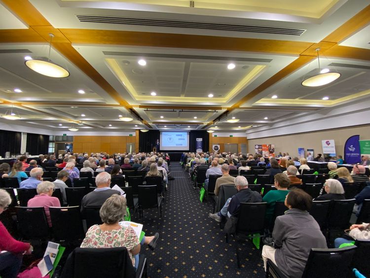 Audience seated in a large conference hall, facing a stage with a presentation screen.