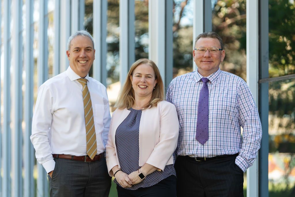 Three professionals smiling, standing outside a modern building. Two men in shirts and ties, one woman in a blazer.