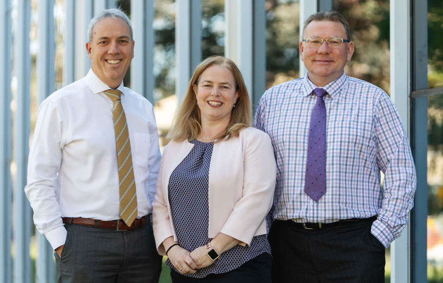 Three professionals standing outdoors: a man with a gold tie, a woman, and a man with a purple tie, smiling.
