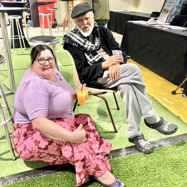 Woman in pink floral skirt and man in hat sit on green turf with drinks.