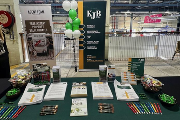 A display table at an event, with KJB branding, marketing materials, pens, and green/white balloons.