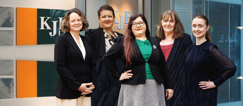 Five women in business attire stand in front of a logo. They are smiling with arms akimbo.