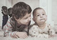 Two young children lying on a fluffy white rug. Older child looks at the baby, who gazes upwards.