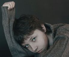 A young person with blue eyes and dark hair, resting their head on an arm, looking pensive.