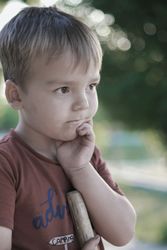 Boy with thoughtful expression, hand on chin, holding a wooden object outdoors.