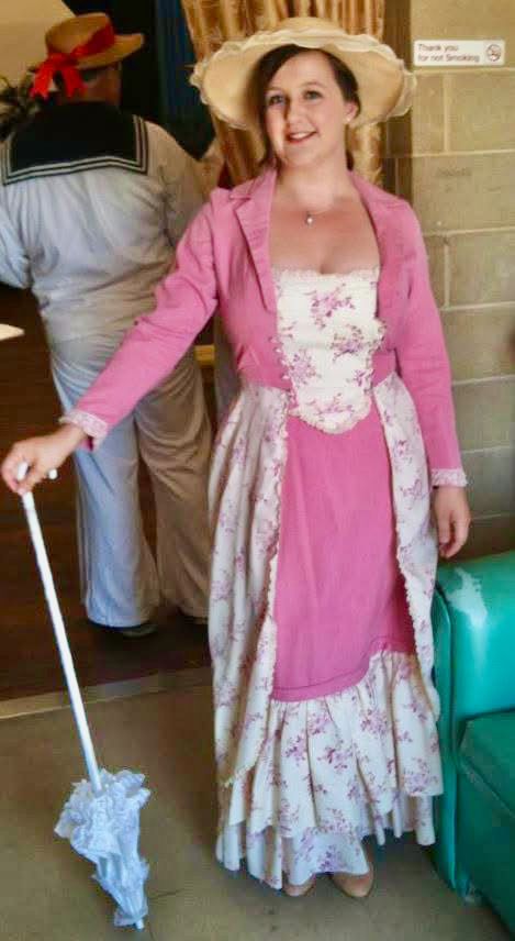 Woman in pink historical costume holding a parasol, smiling. Another person in a sailor outfit in the background.