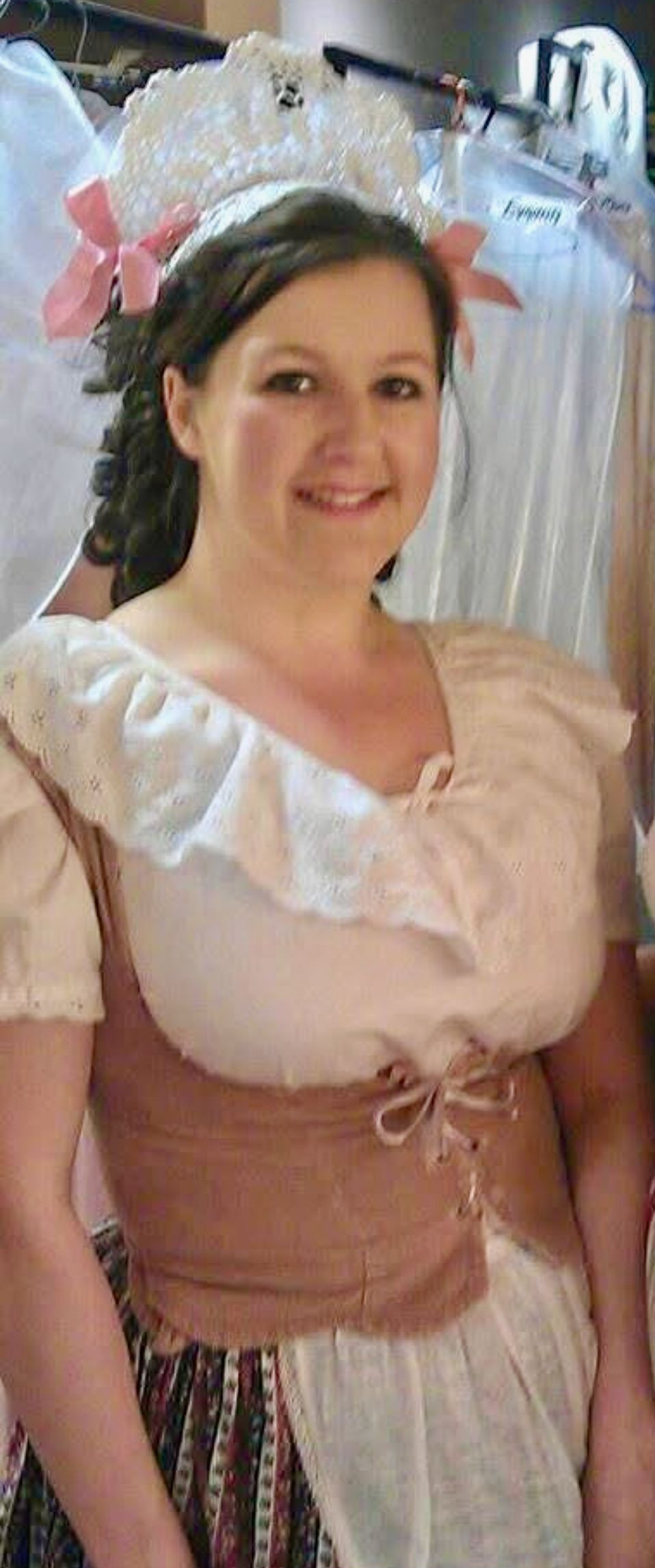 Woman in period costume smiles. She wears a headpiece with pink bows, a white blouse with ruffles, and a brown corset.