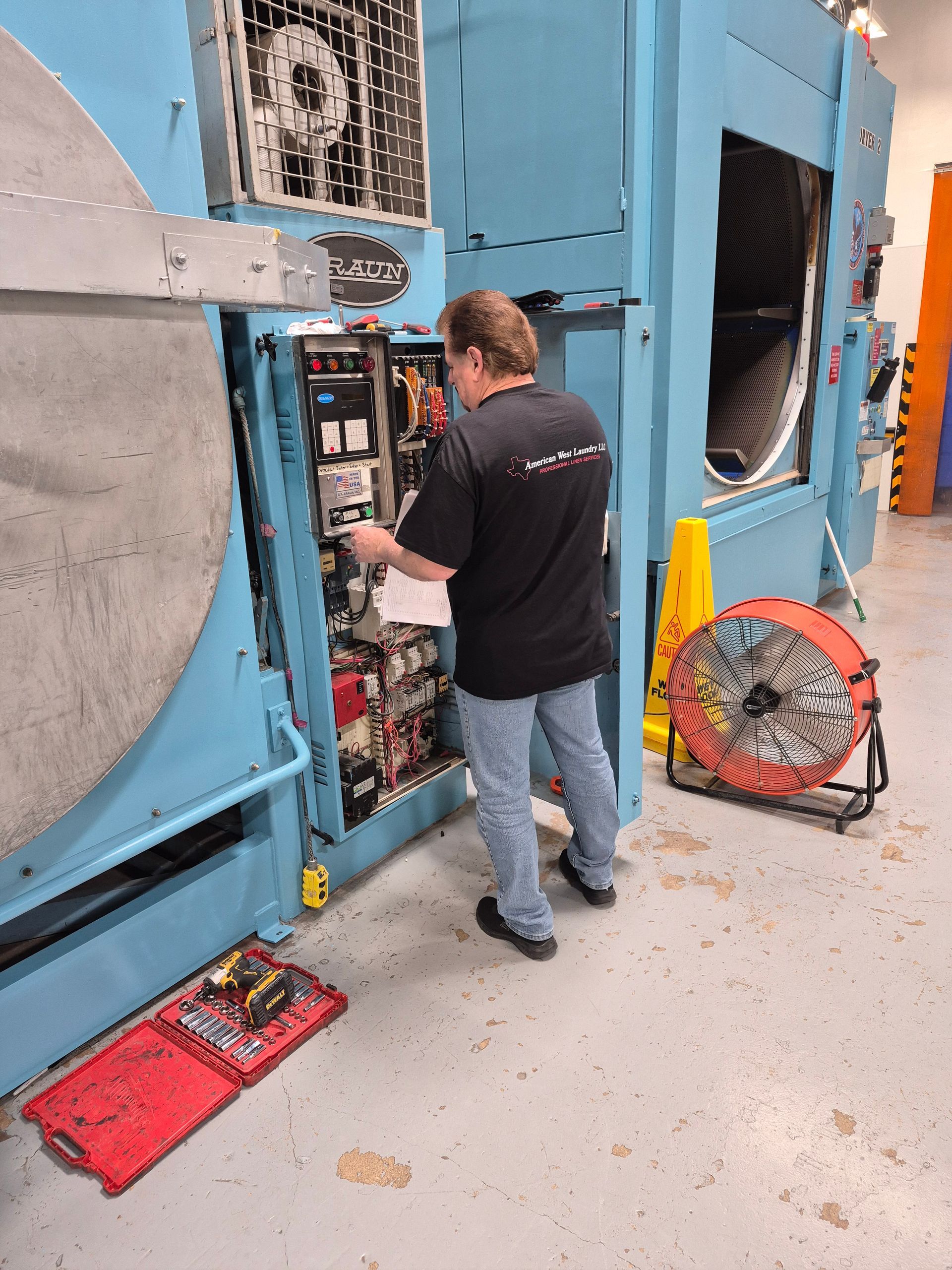 Man in black shirt repairs a large, blue machine. He's working on an electrical panel, tools on the floor.