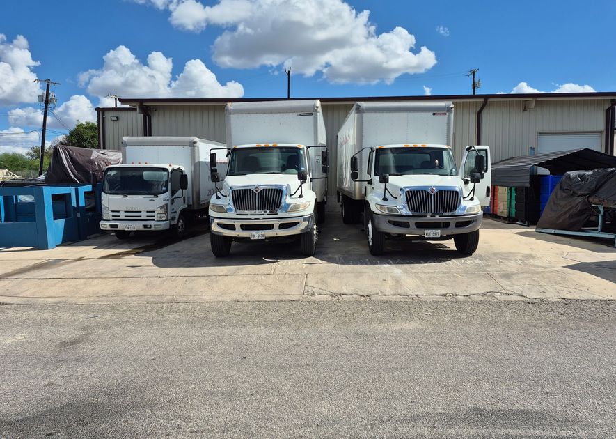 Three white box trucks parked in front of a building on a sunny day.