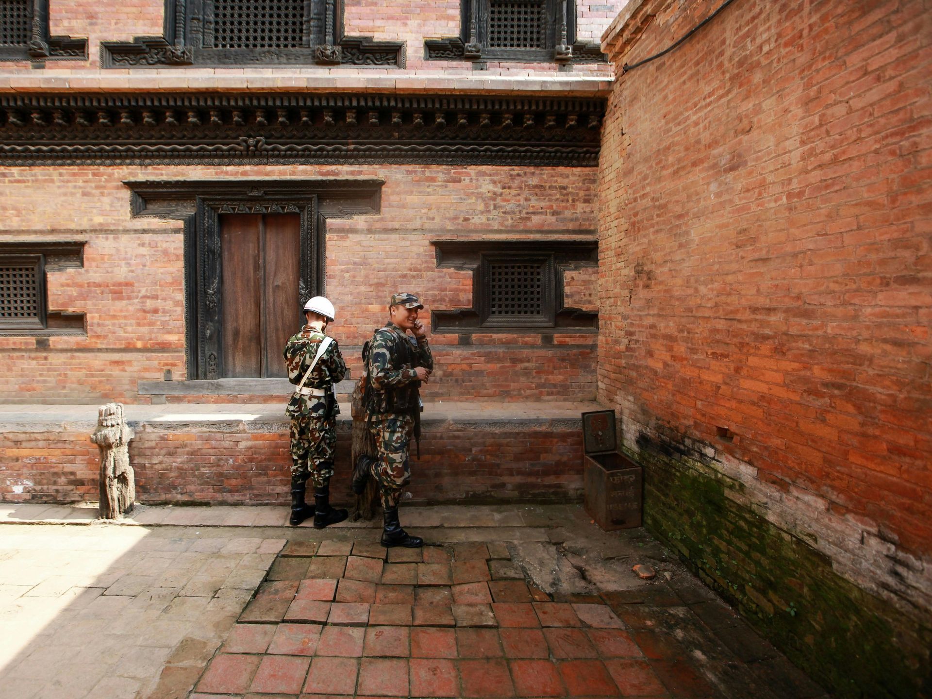 Two soldiers in camouflage uniform stand guard near a red brick building in Nepal.