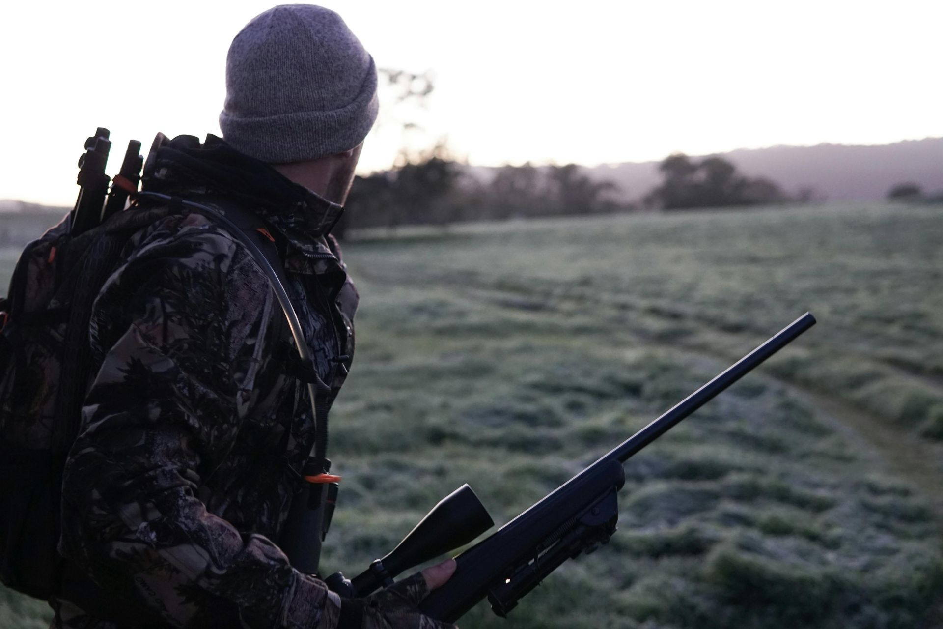 Hunter in camouflage gear, holding rifle, looking at a field in the early morning.