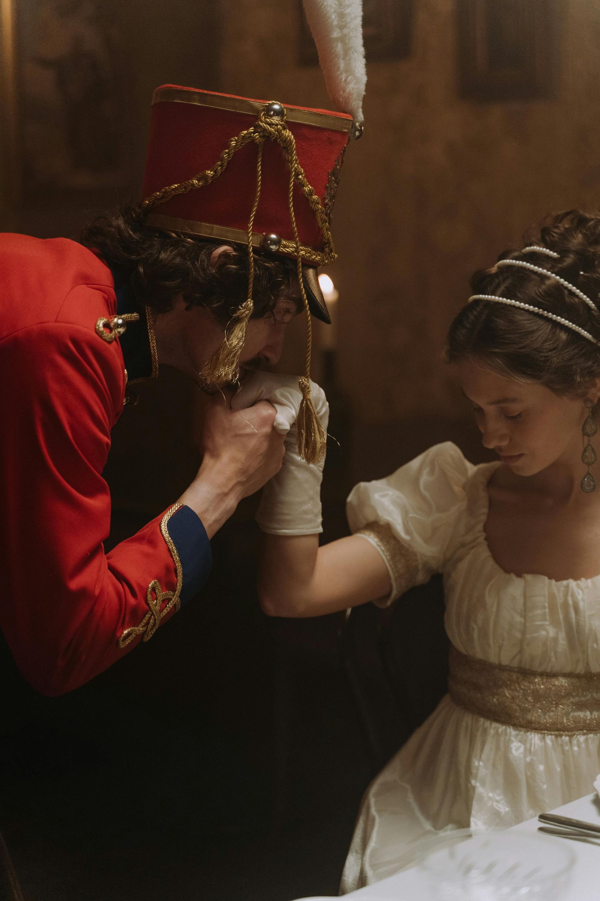 A man in a red military jacket kisses a gloved hand of a young woman in a white dress.