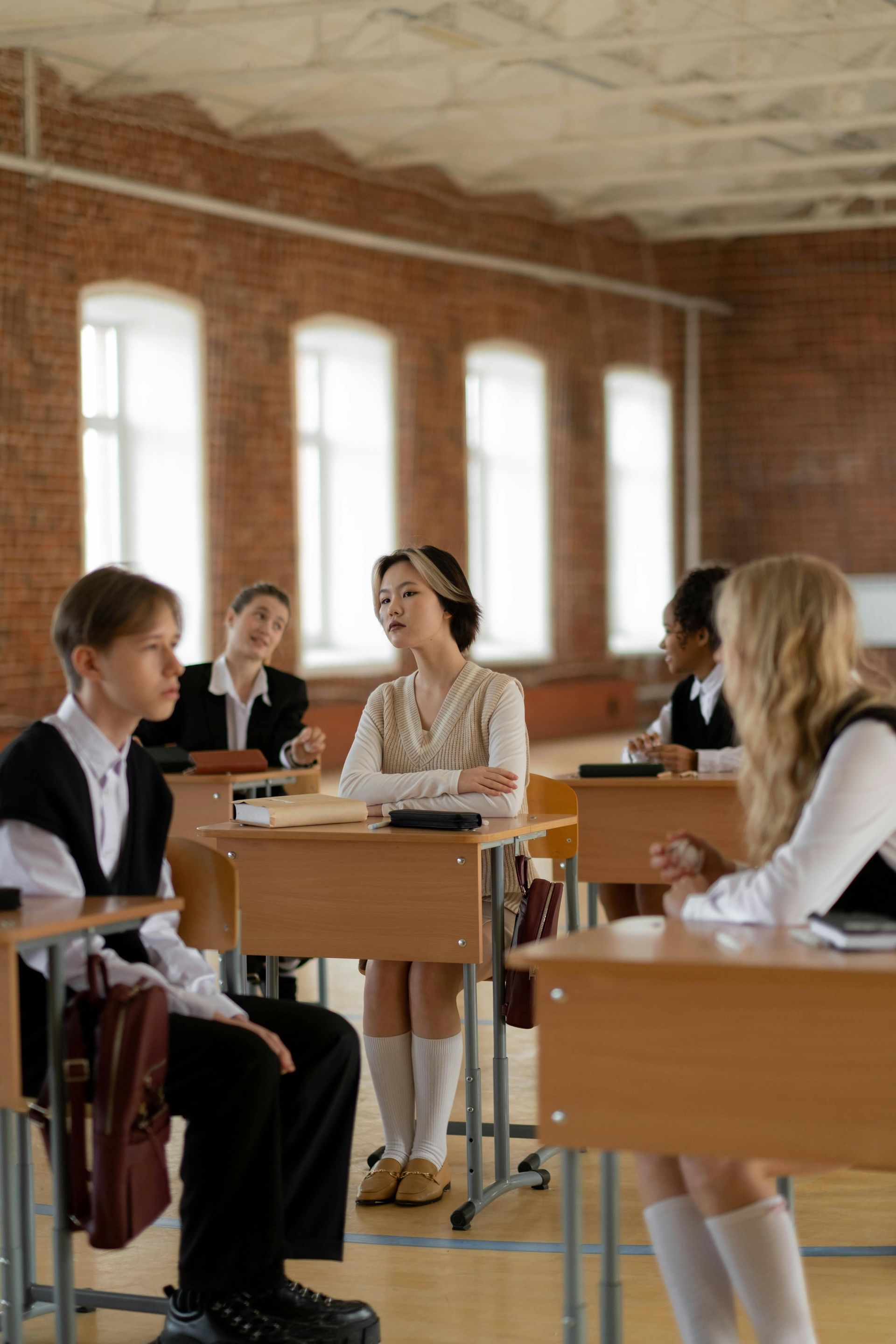 Students in a brick-walled classroom. One student speaks, while others listen. Desks, windows, and a formal dress code are visible.