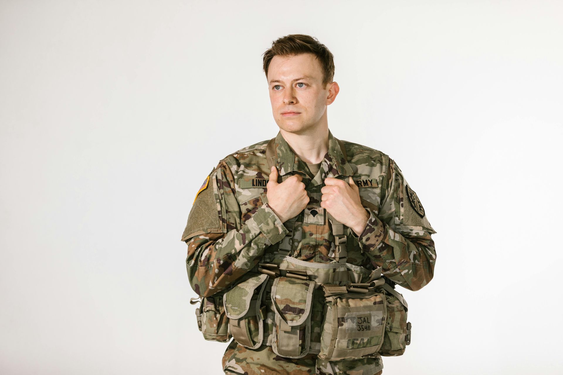 Soldier in camouflage uniform looking to the side, hands on chest, against a white backdrop.