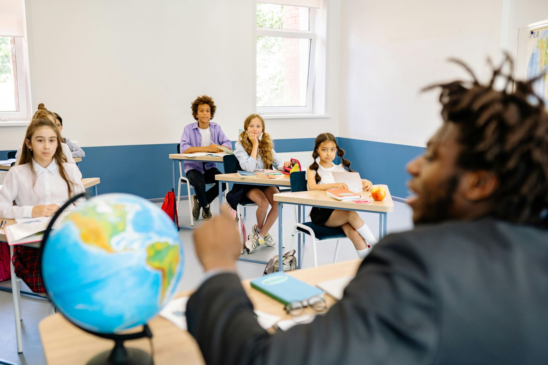 Teacher points at globe in classroom, students at desks, white walls, blue trim.