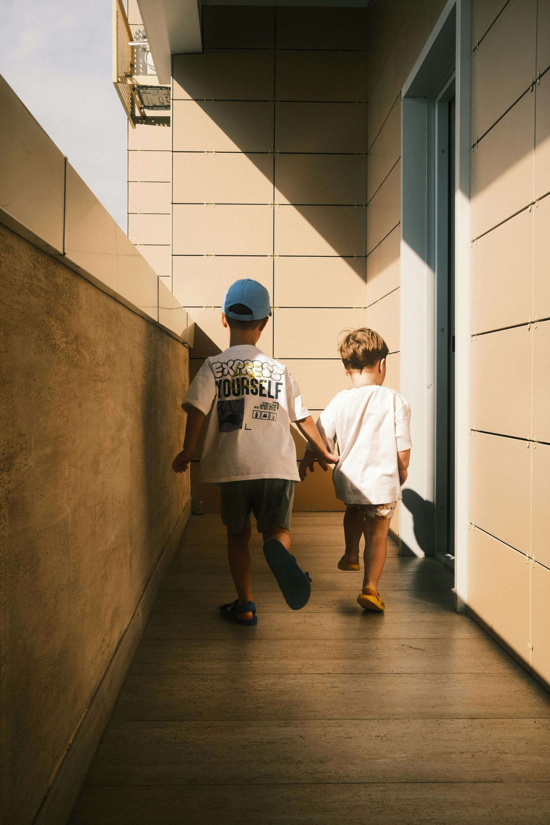 Two children holding hands walk down a sunlit hallway. One wears a blue hat, the other has curly hair.