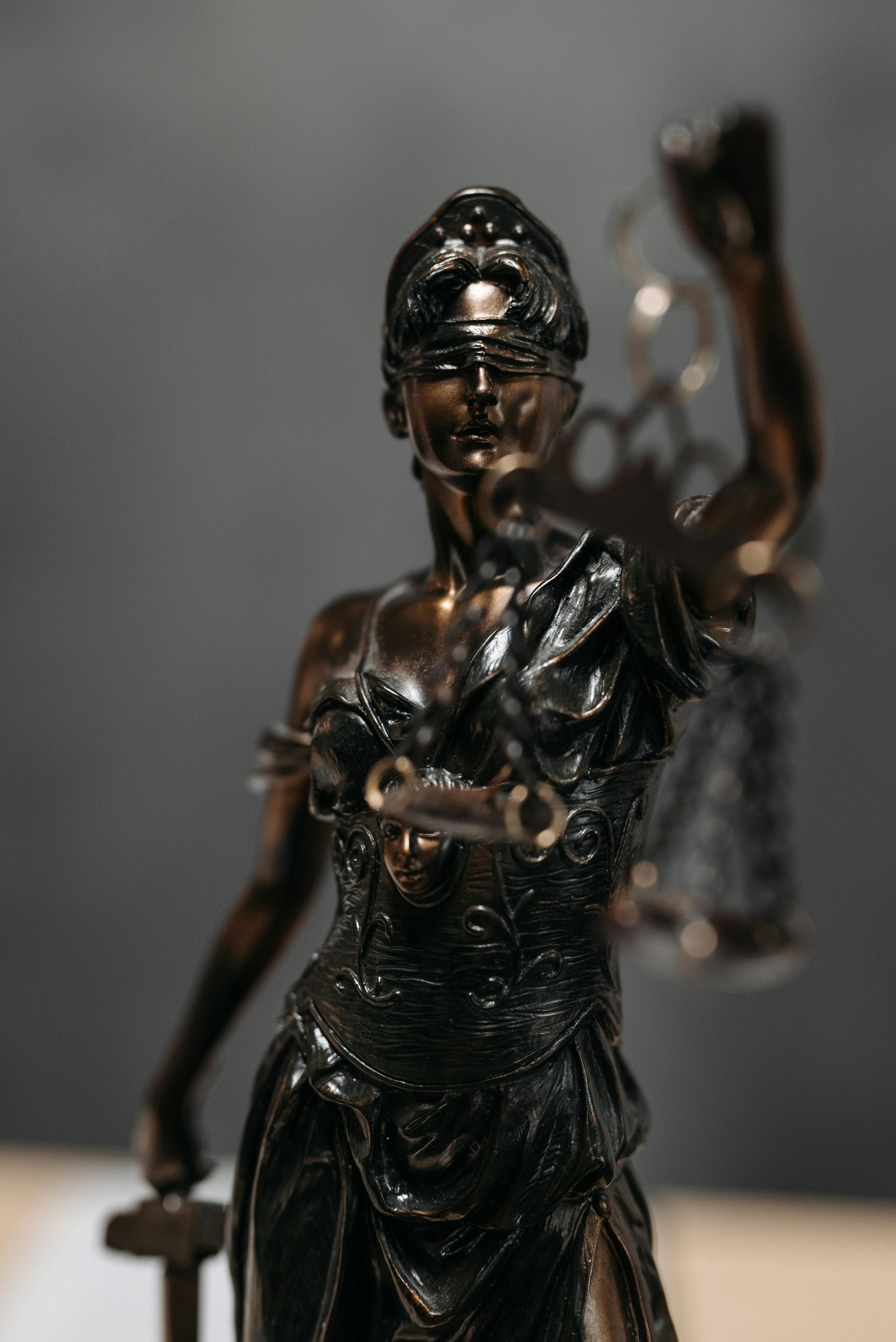 Statue of Lady Justice with blindfold, holding scales and sword. Bronze color against gray background.