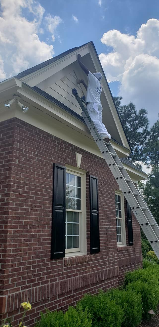 Person in protective suit on a ladder, painting the trim of a brick house.