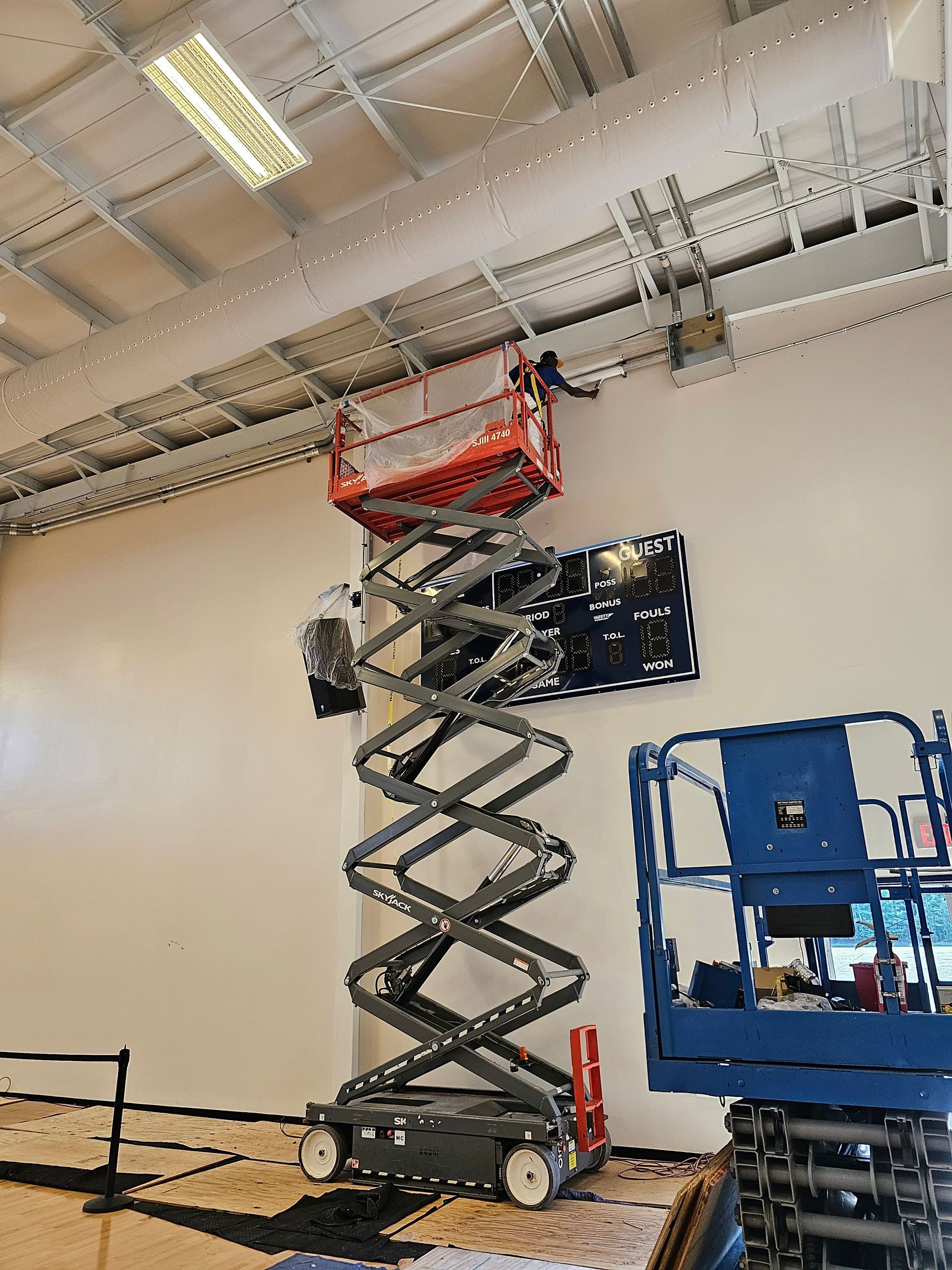 A scissor lift elevates a worker to reach ceiling-mounted conduits in a commercial space with a white wall.