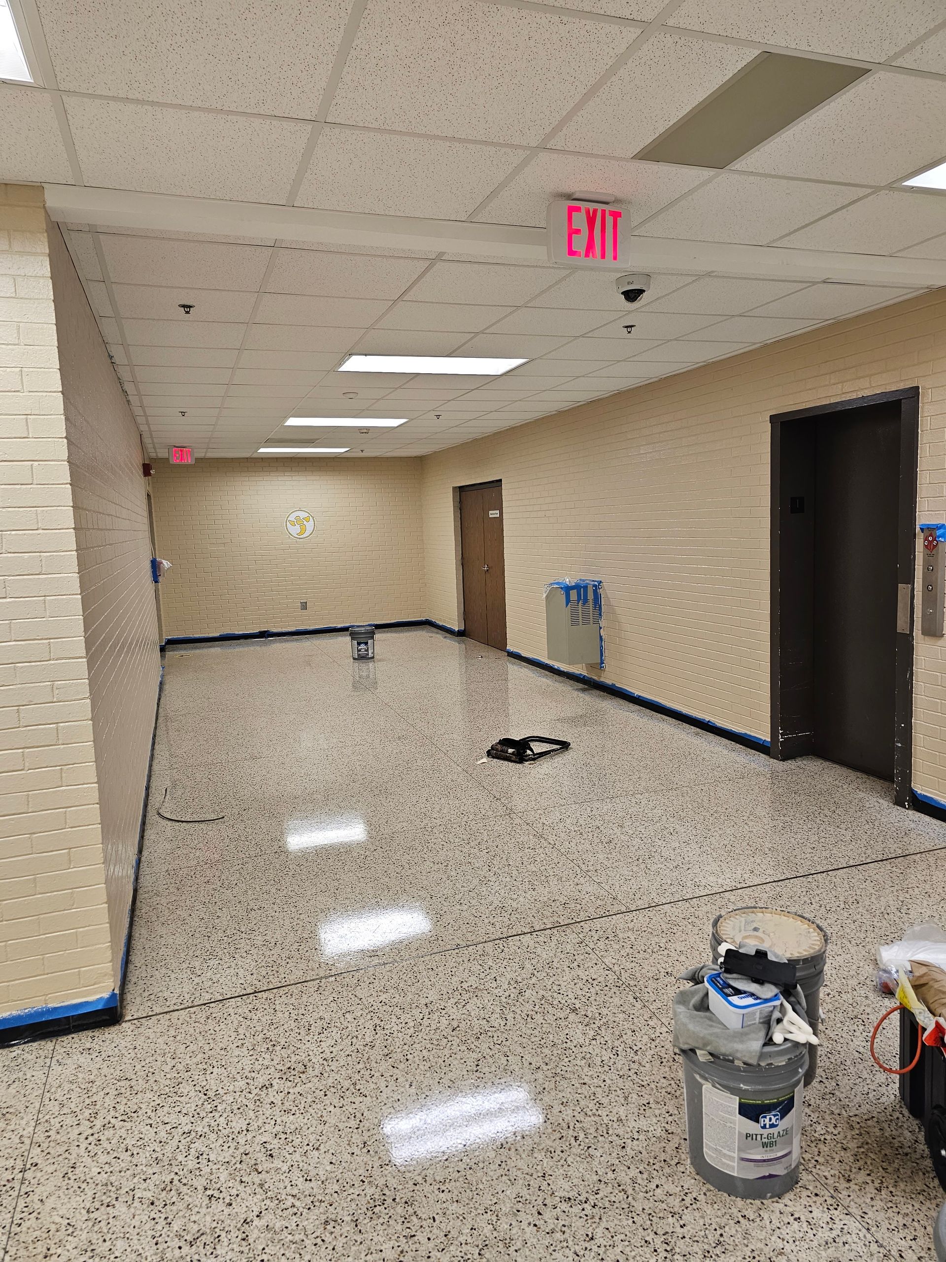 Hallway with speckled floor, tan walls, exit sign, and painting supplies.