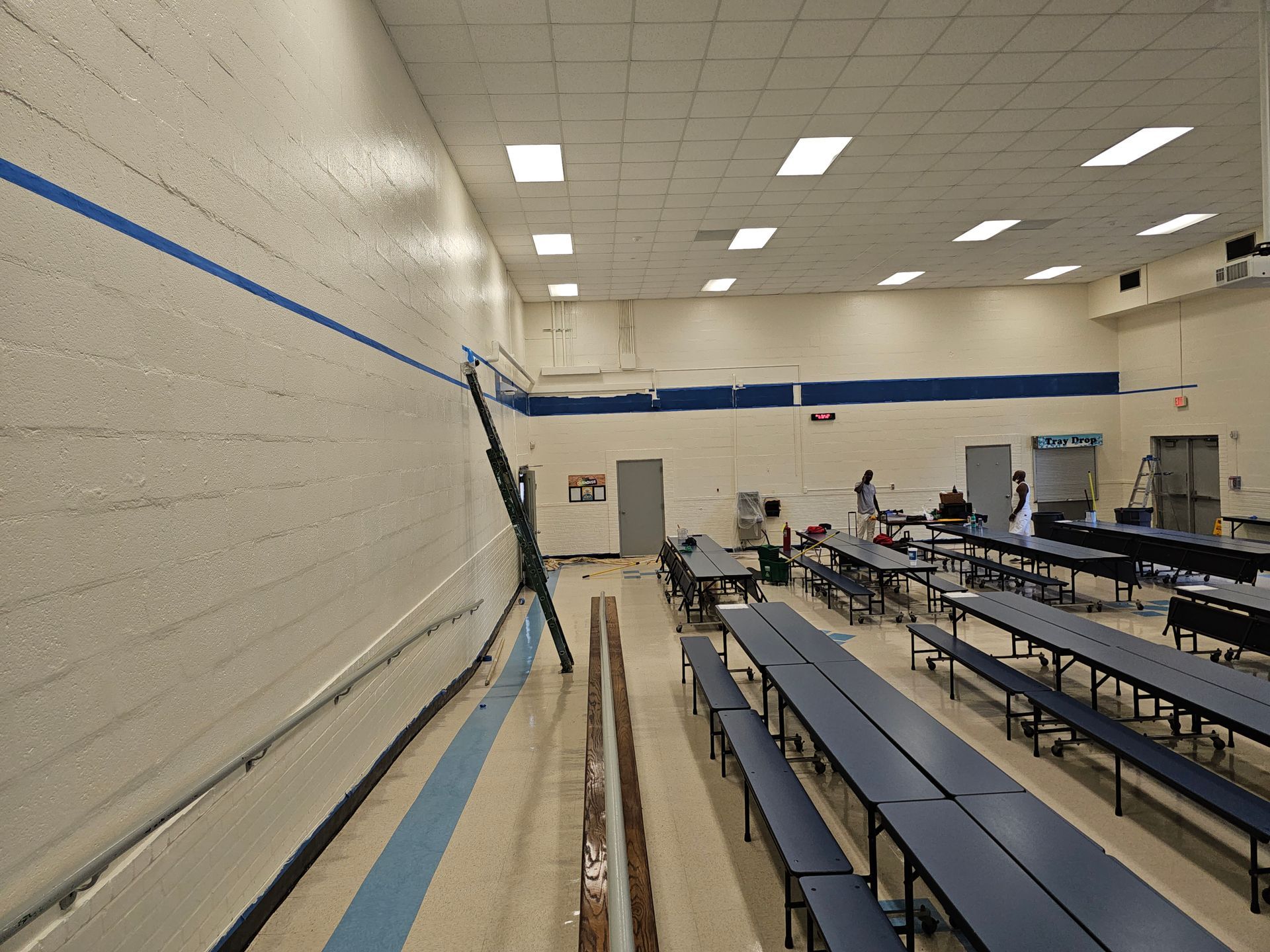 A school cafeteria with long blue tables and a damaged wall.