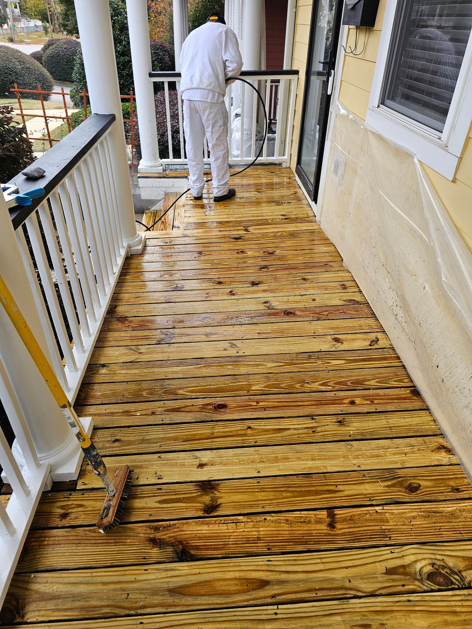 Person pressure washing a wooden porch, cleaning the planks, surrounded by railings and a yellow wall.