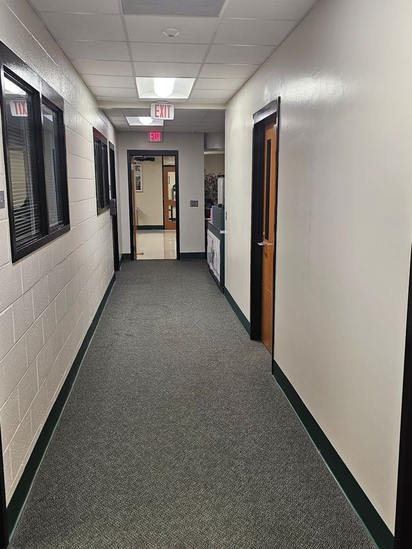 A long hallway with carpet, doors, and windows. A door with an exit sign at the end of the hall.