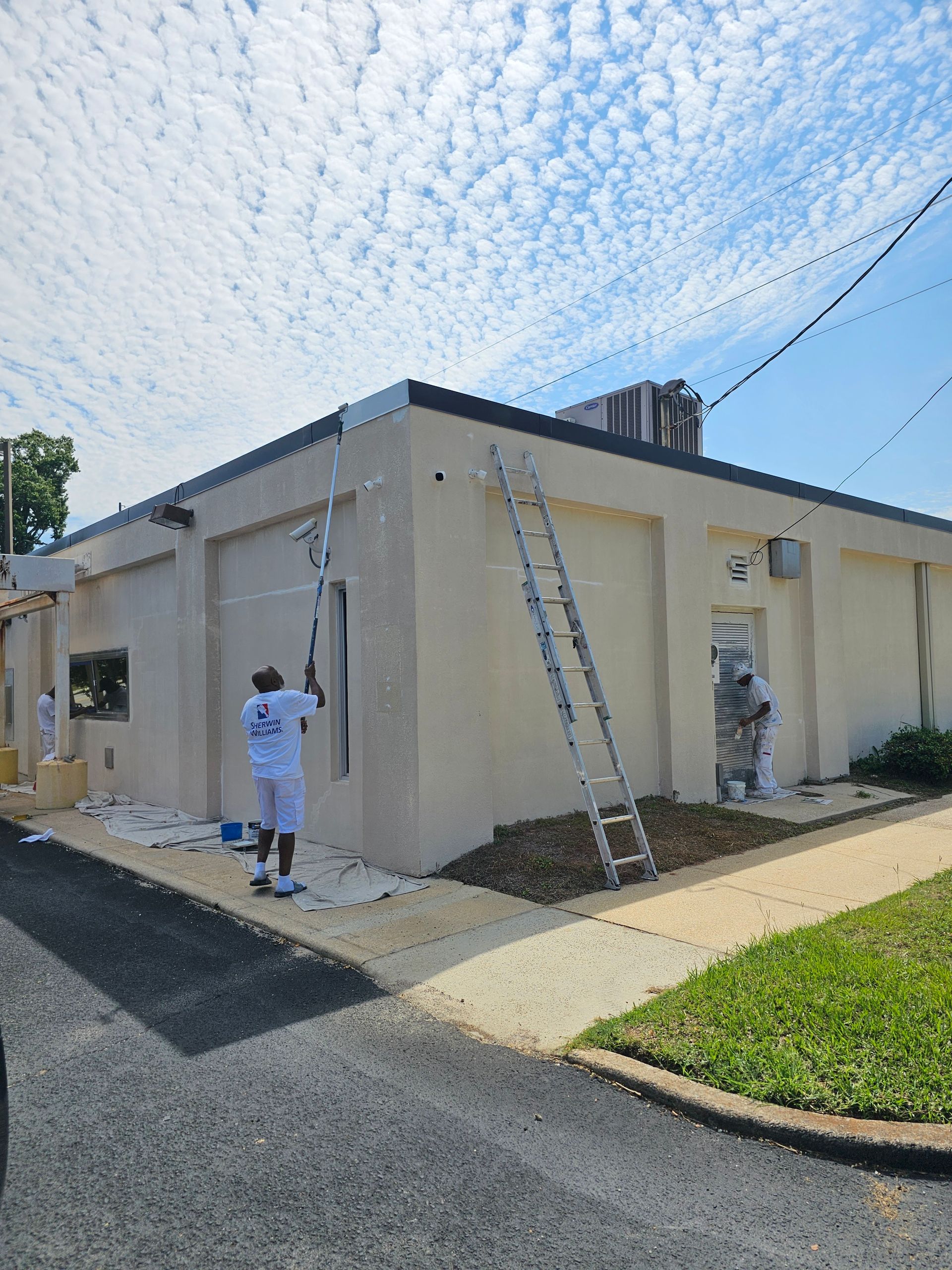 Two people painting a tan building exterior with a ladder and roller under a partly cloudy sky.