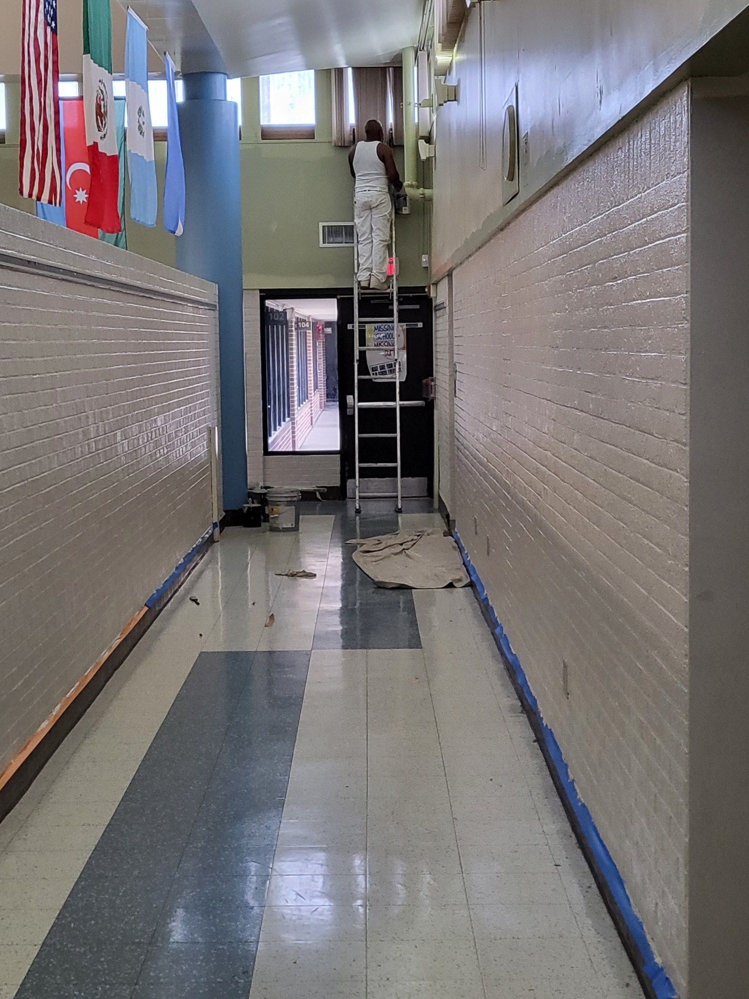 Person on ladder painting a school hallway. Blue tape on the floor, flags in the background.
