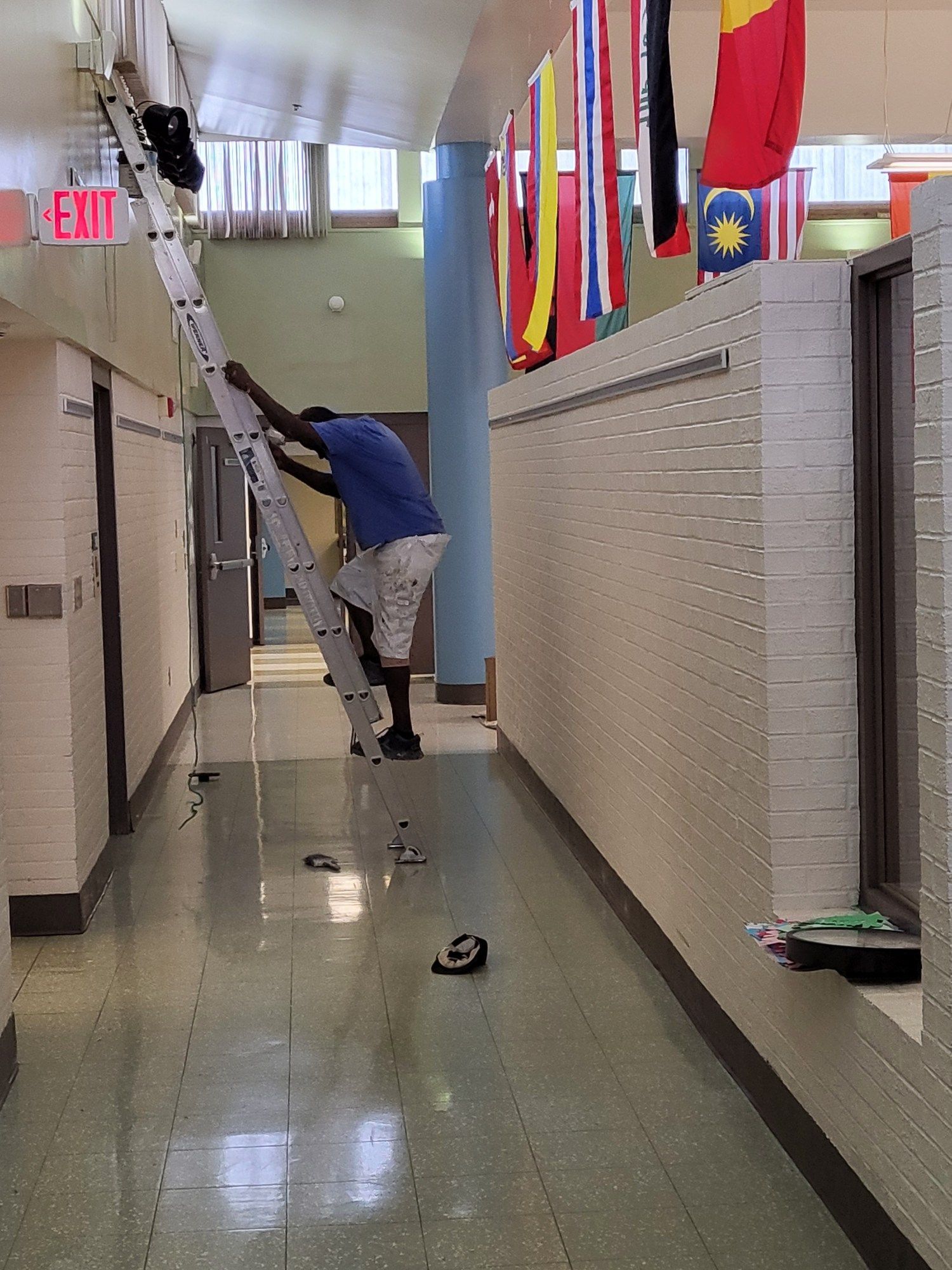 Person climbs a ladder in a hallway, reaching toward ceiling.  Hallway has flags and an exit sign.