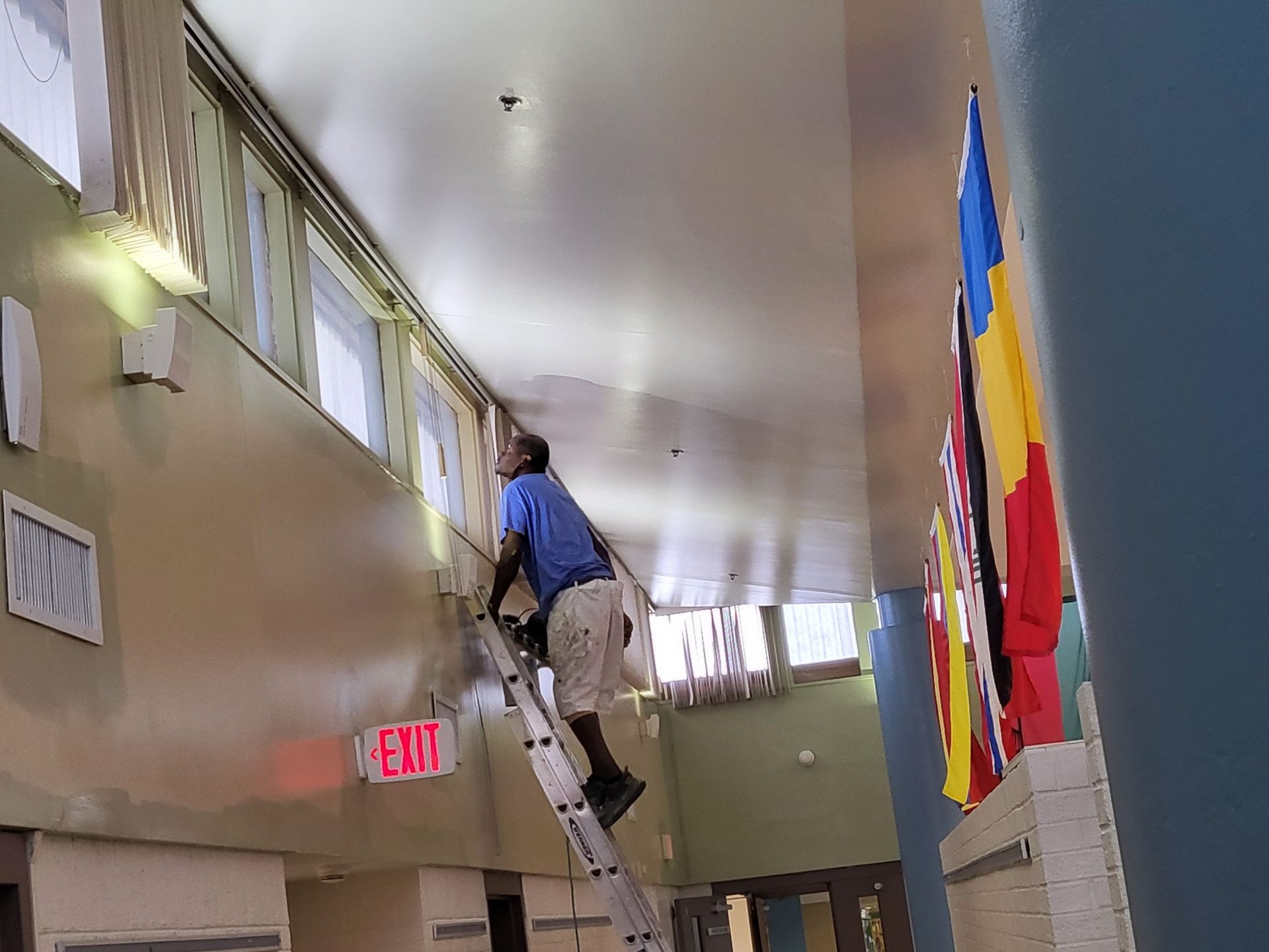 A person on a ladder paints a ceiling in a building's interior. 