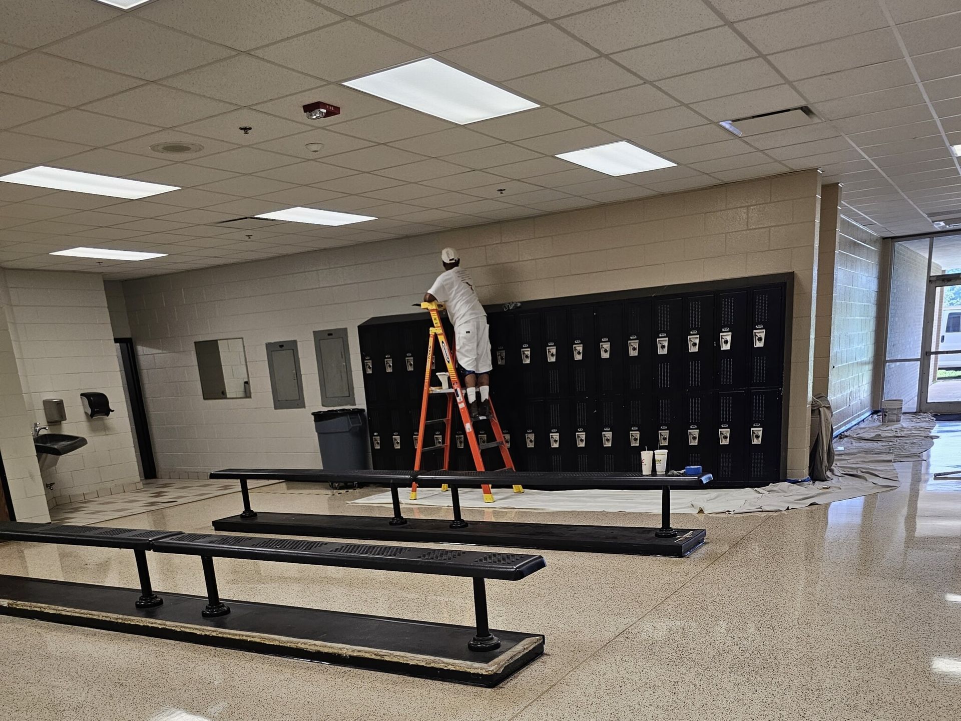 Person on ladder painting a wall above black lockers in a school hallway.