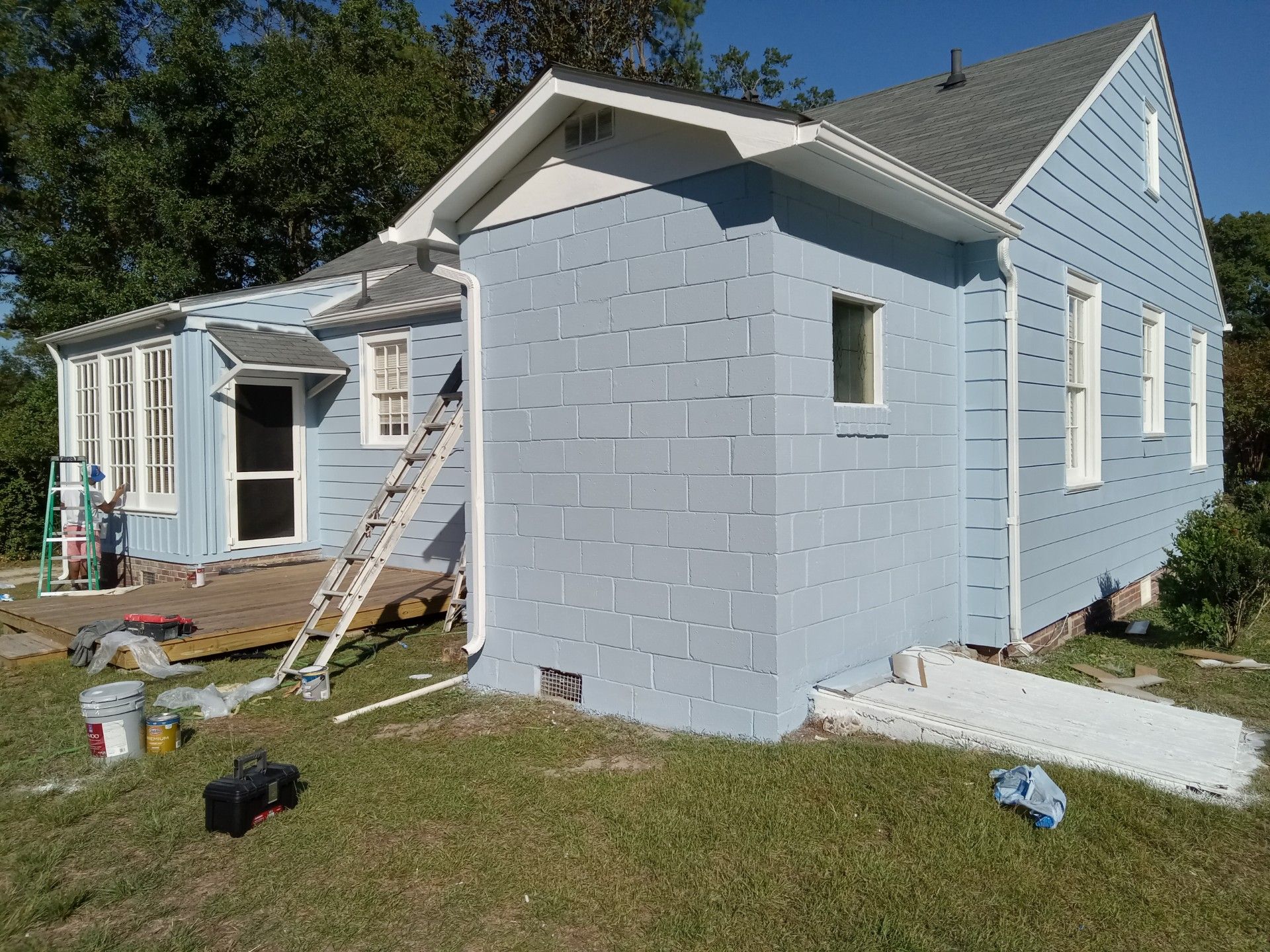 Light blue house with white trim being painted. Ladder, paint cans, and supplies are visible.