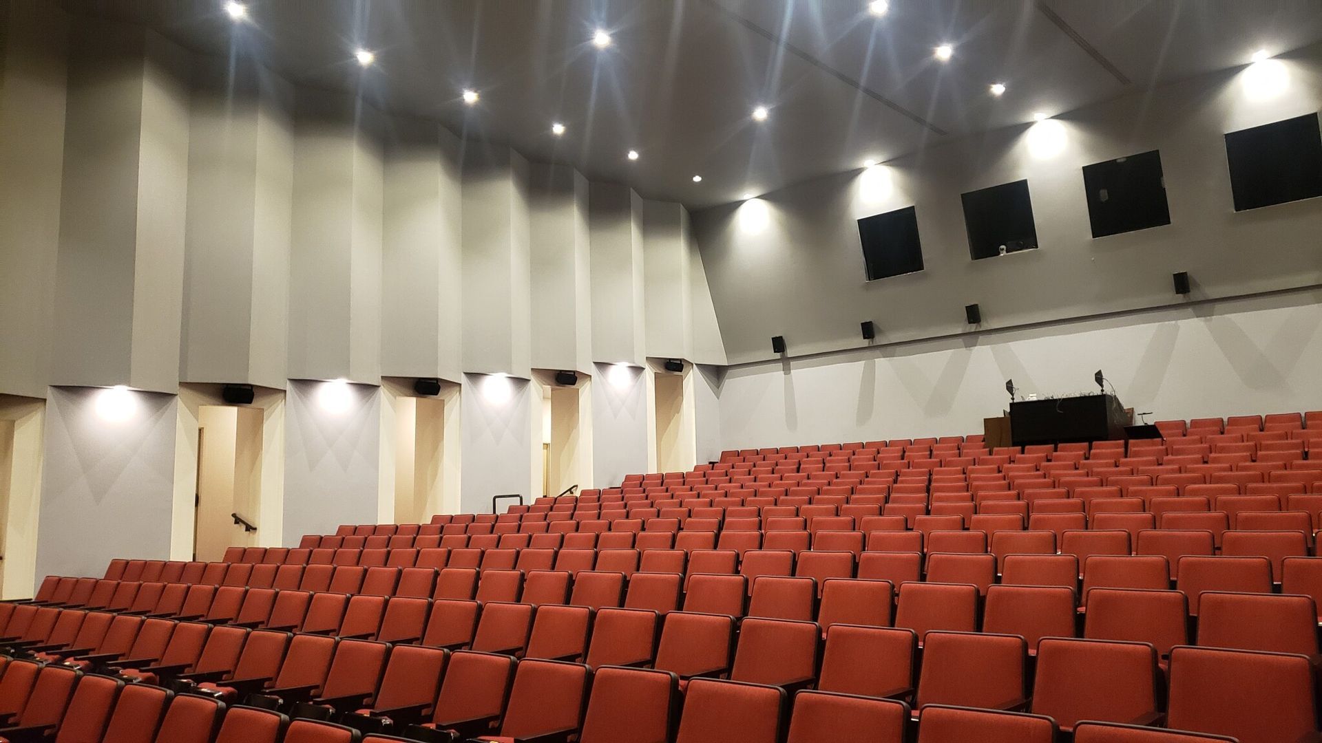 Auditorium with rows of red seats, stage at the end, and recessed lighting on the ceiling.