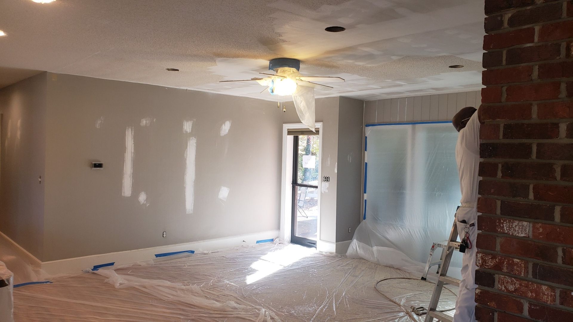 Room being painted: gray walls, protective plastic on floor, painter on ladder by taped-off doorway, and brick wall.