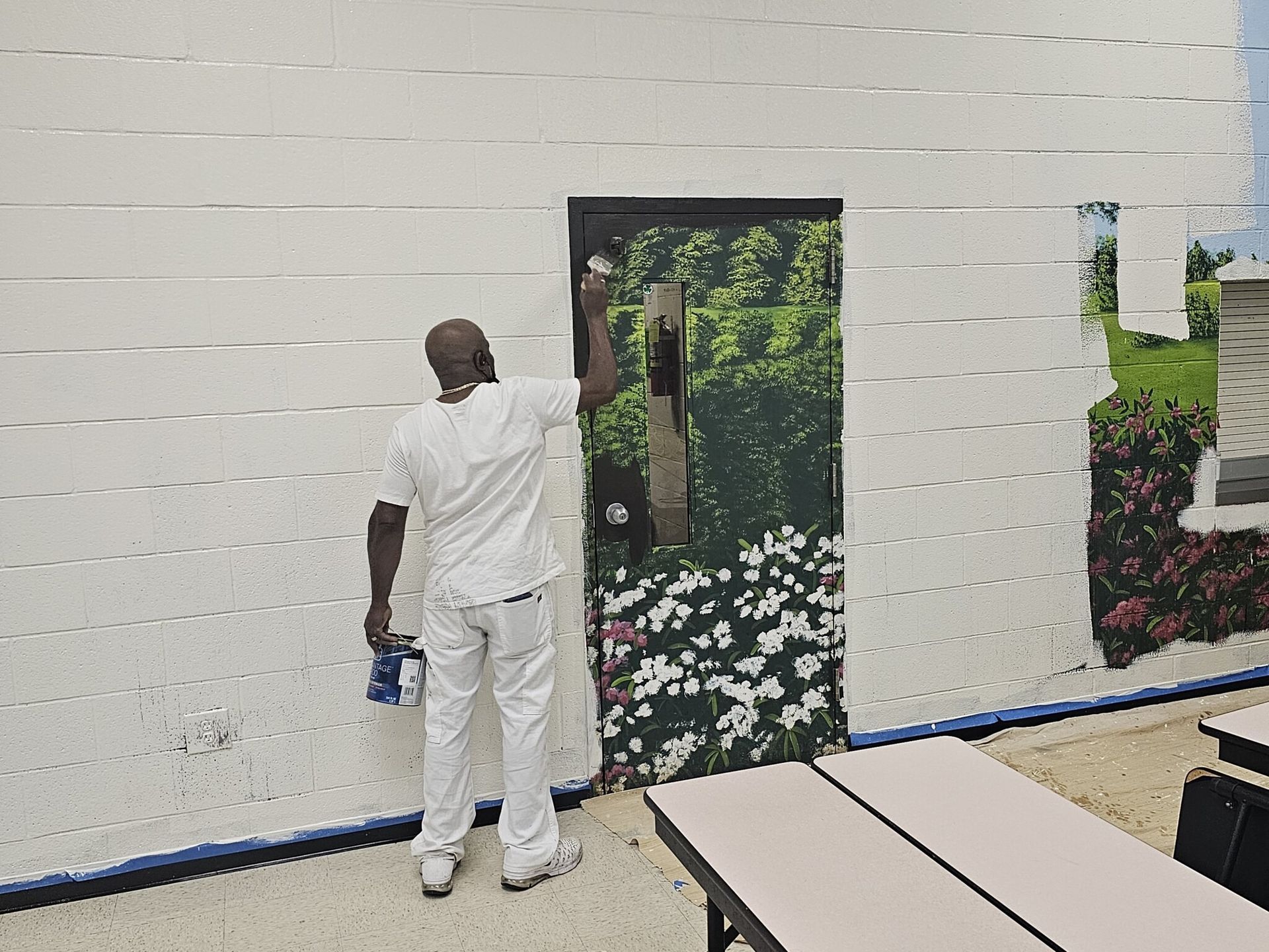 Man in white painting a mural of a doorway with a garden view on a white wall.