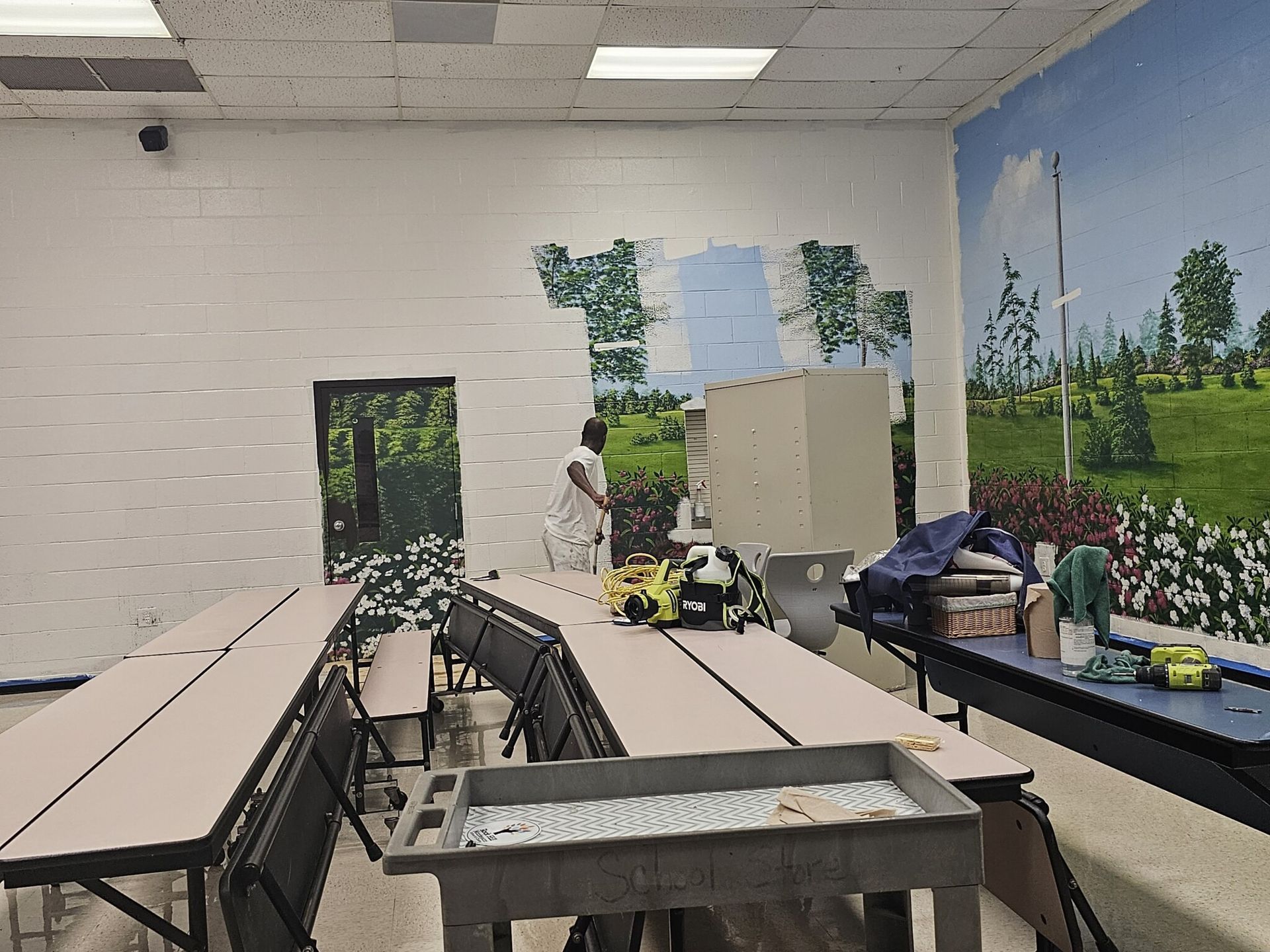 A person painting a mural in a school cafeteria. Picnic tables and a food service counter are also visible.