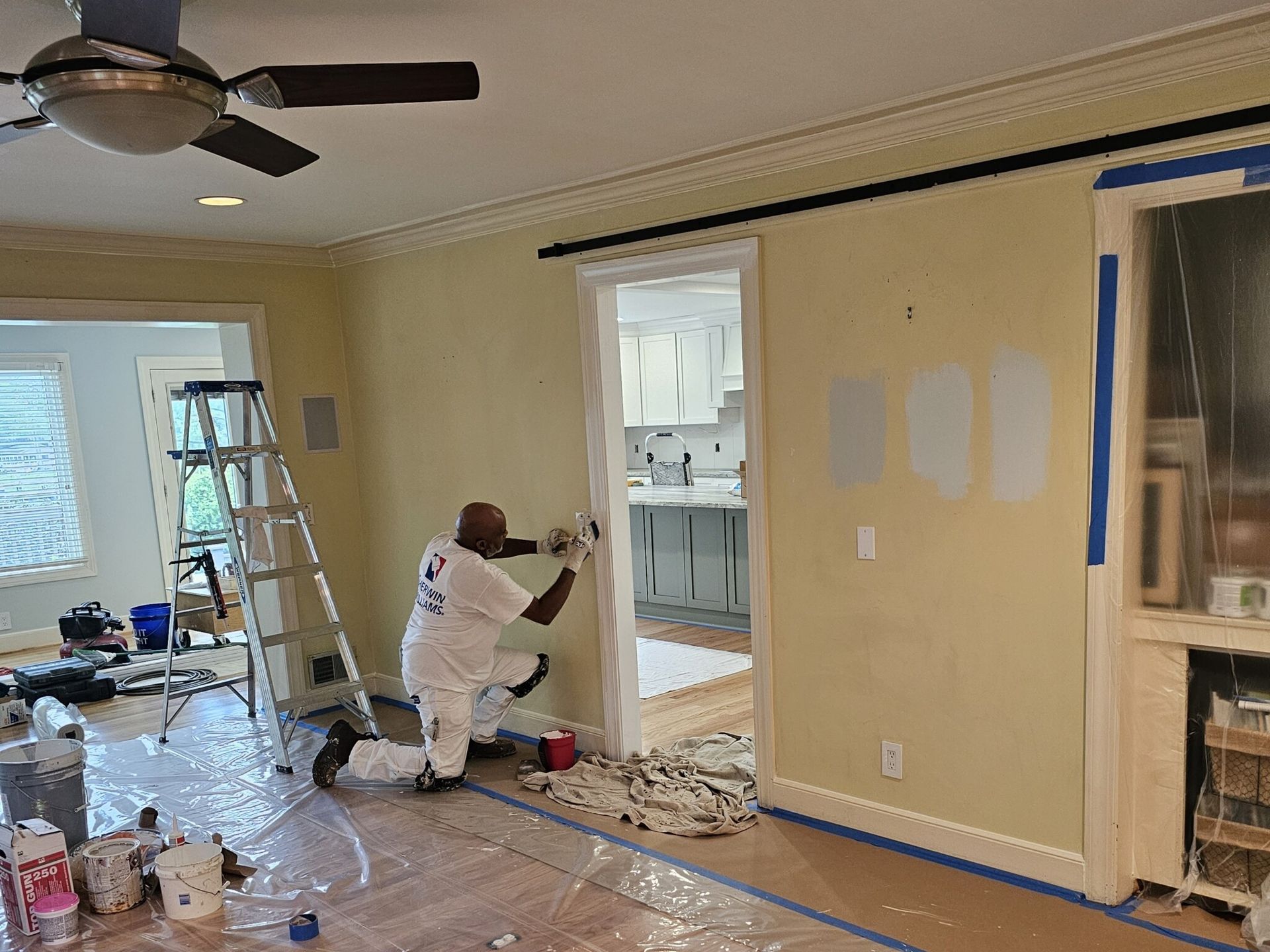 Man painting an interior door frame in a room with taped walls, paint swatches, and plastic sheeting.