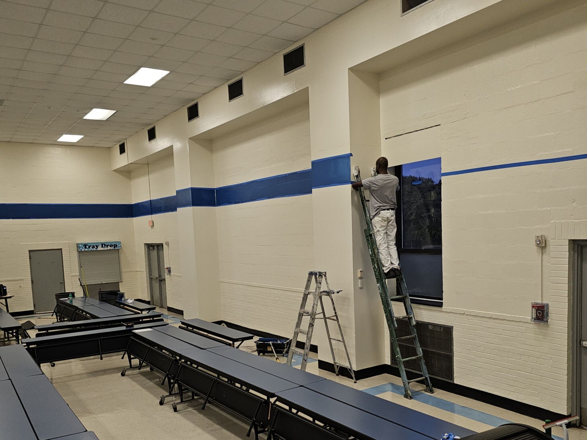Person on ladder painting a wall in a school cafeteria with long tables. Blue and white color scheme.