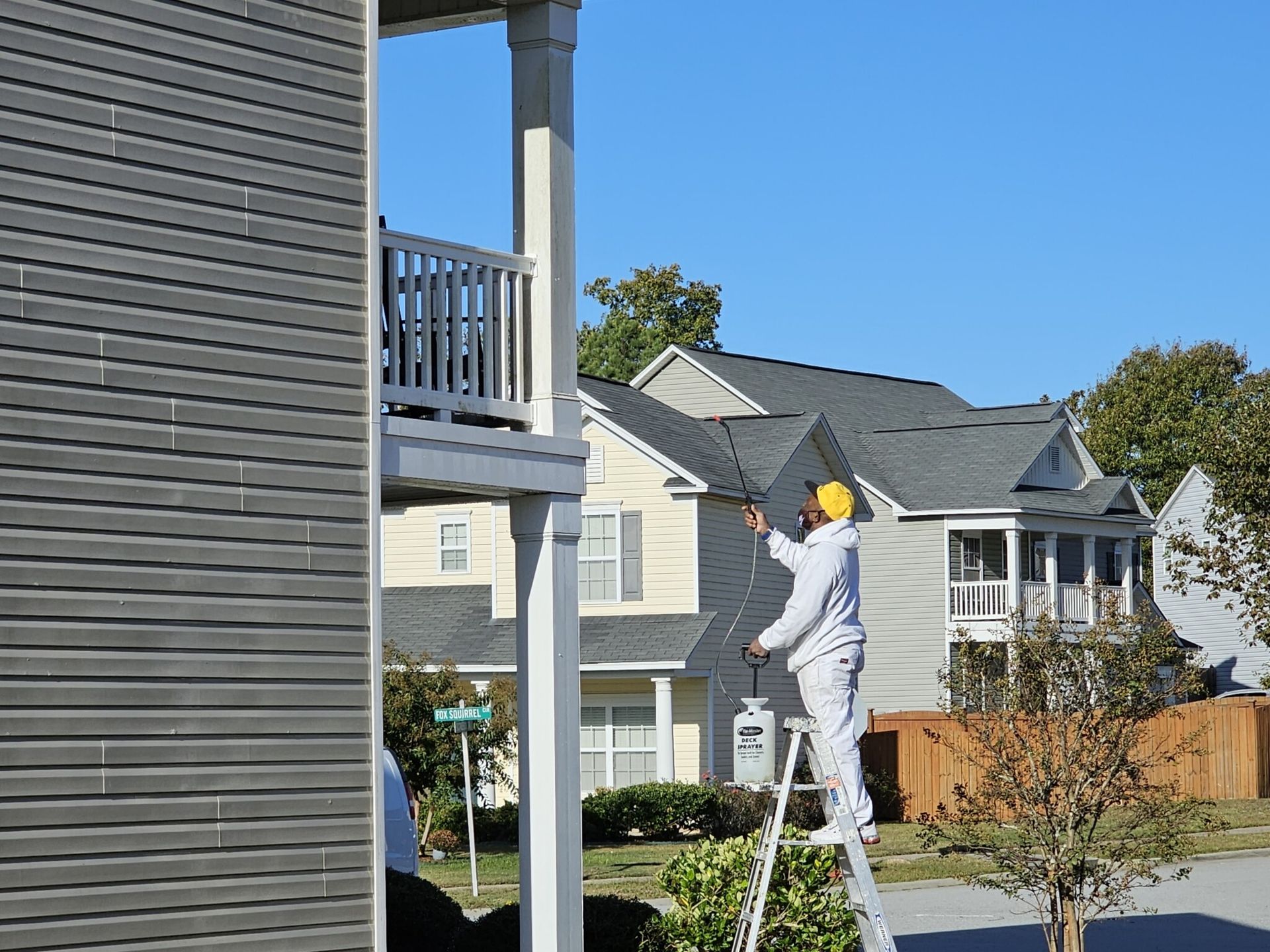 Person painting a house exterior from a ladder; wearing safety gear, blue sky, houses in background.