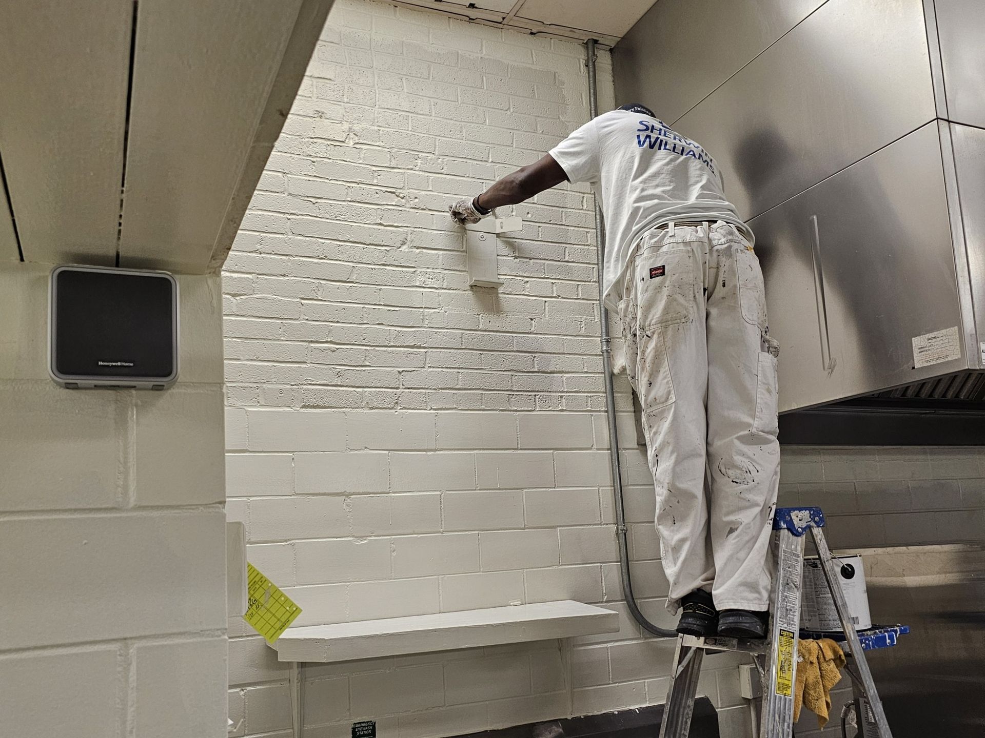Person painting a white brick wall from a ladder in a commercial setting.