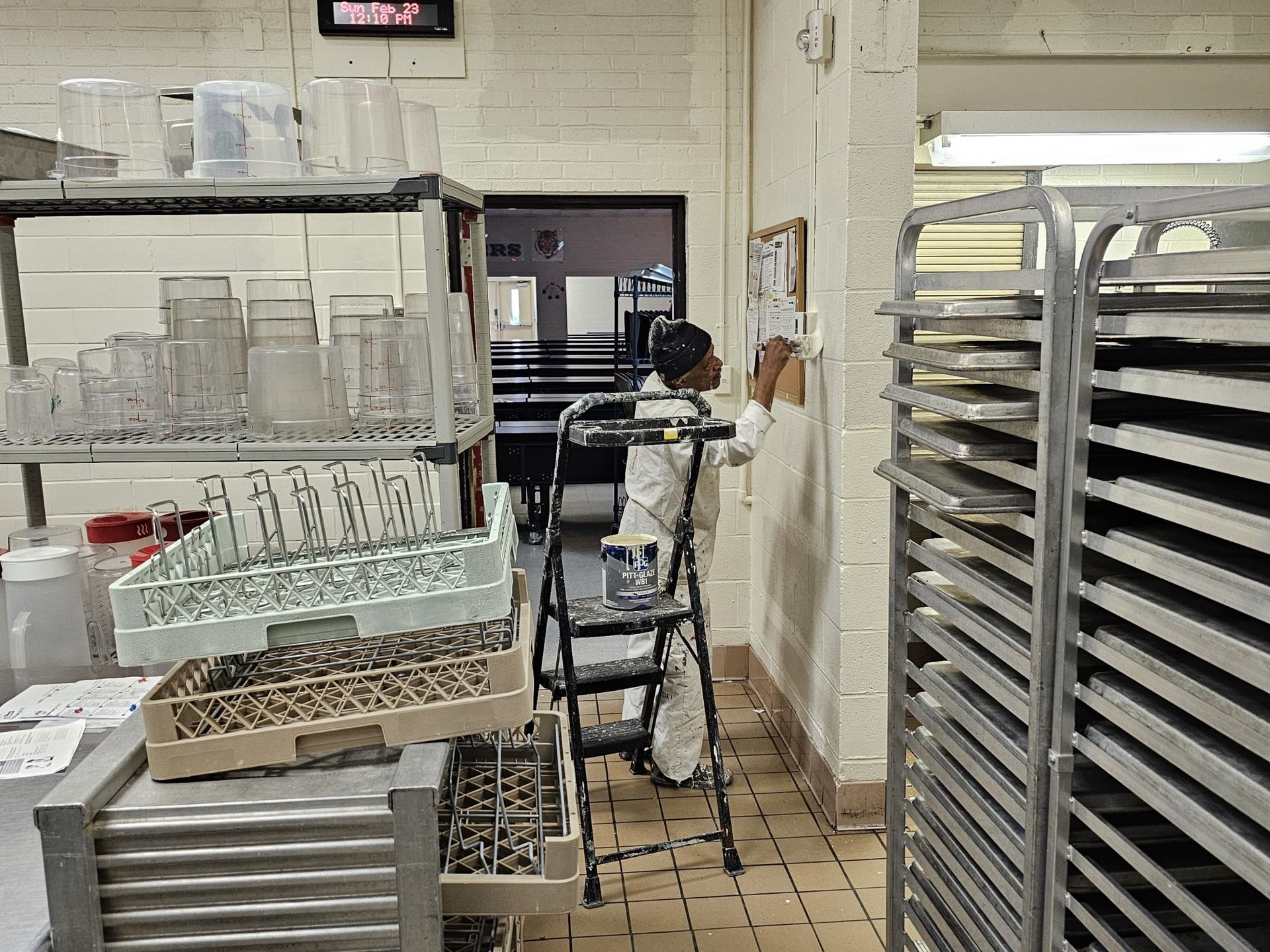 Person on a stepladder in a commercial kitchen, adjusting something on the wall near metal shelving and dish racks.