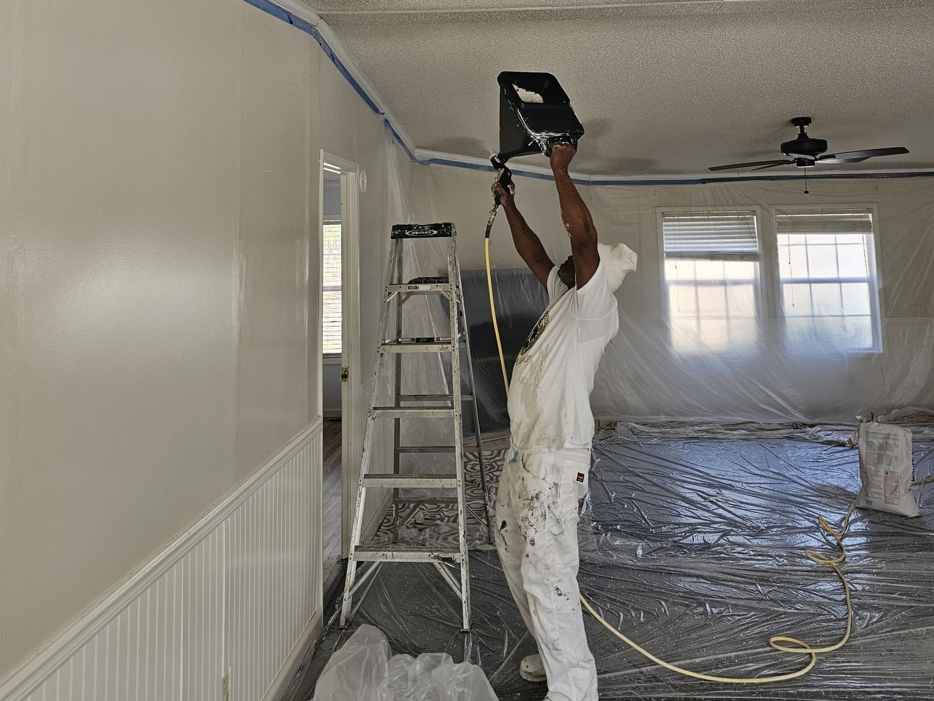 Person painting ceiling, standing on a ladder. Room is partially covered with plastic sheeting; walls are white.