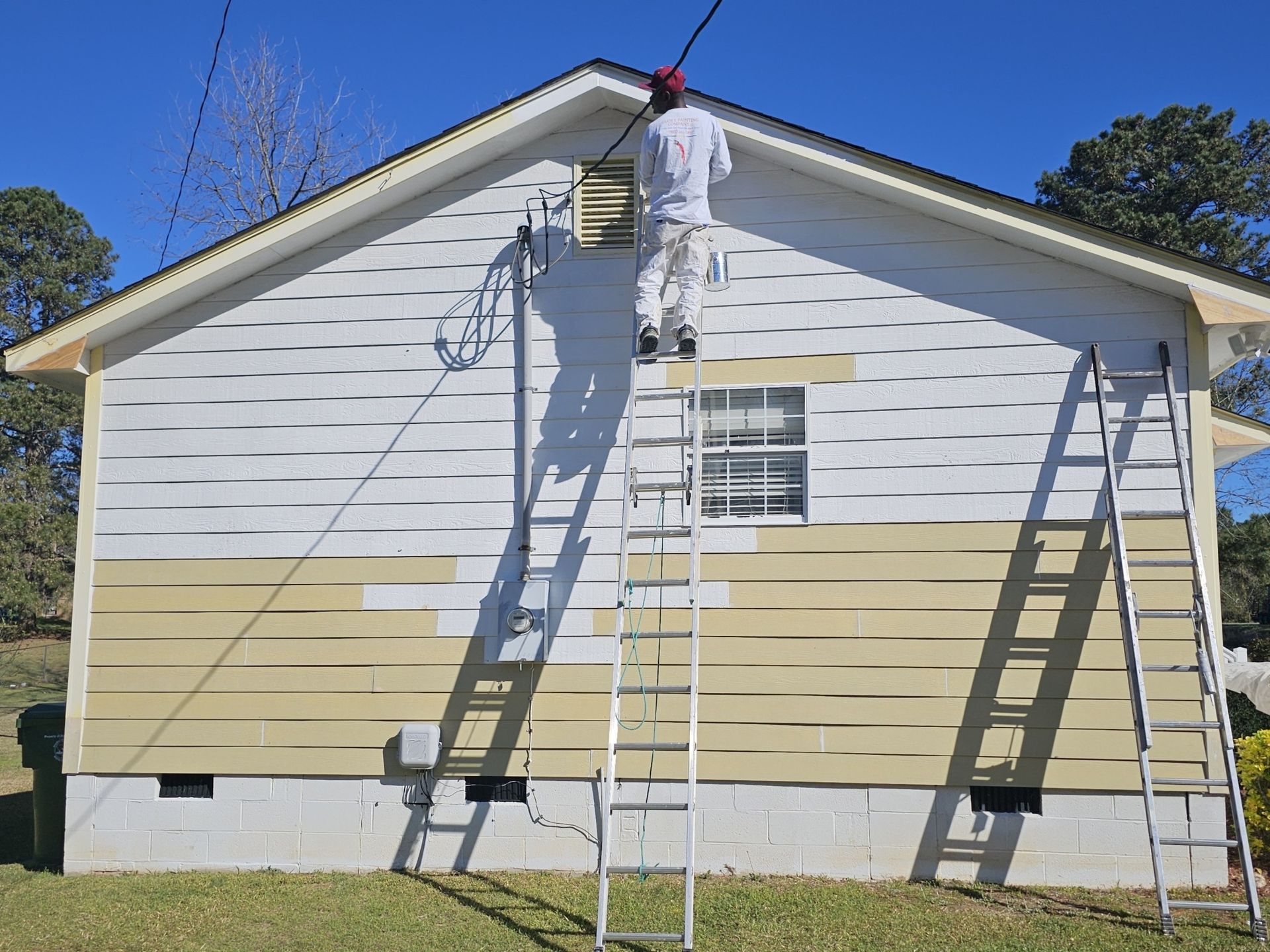 Person on a ladder painting a house. Yellow and white siding with blue sky.