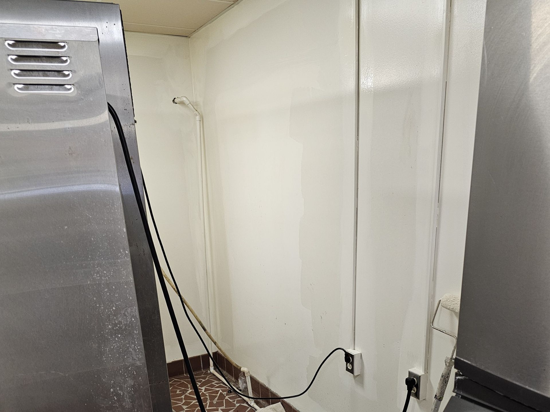 A narrow, white-walled space with electrical outlets, a floor drain, and a stainless steel appliance.