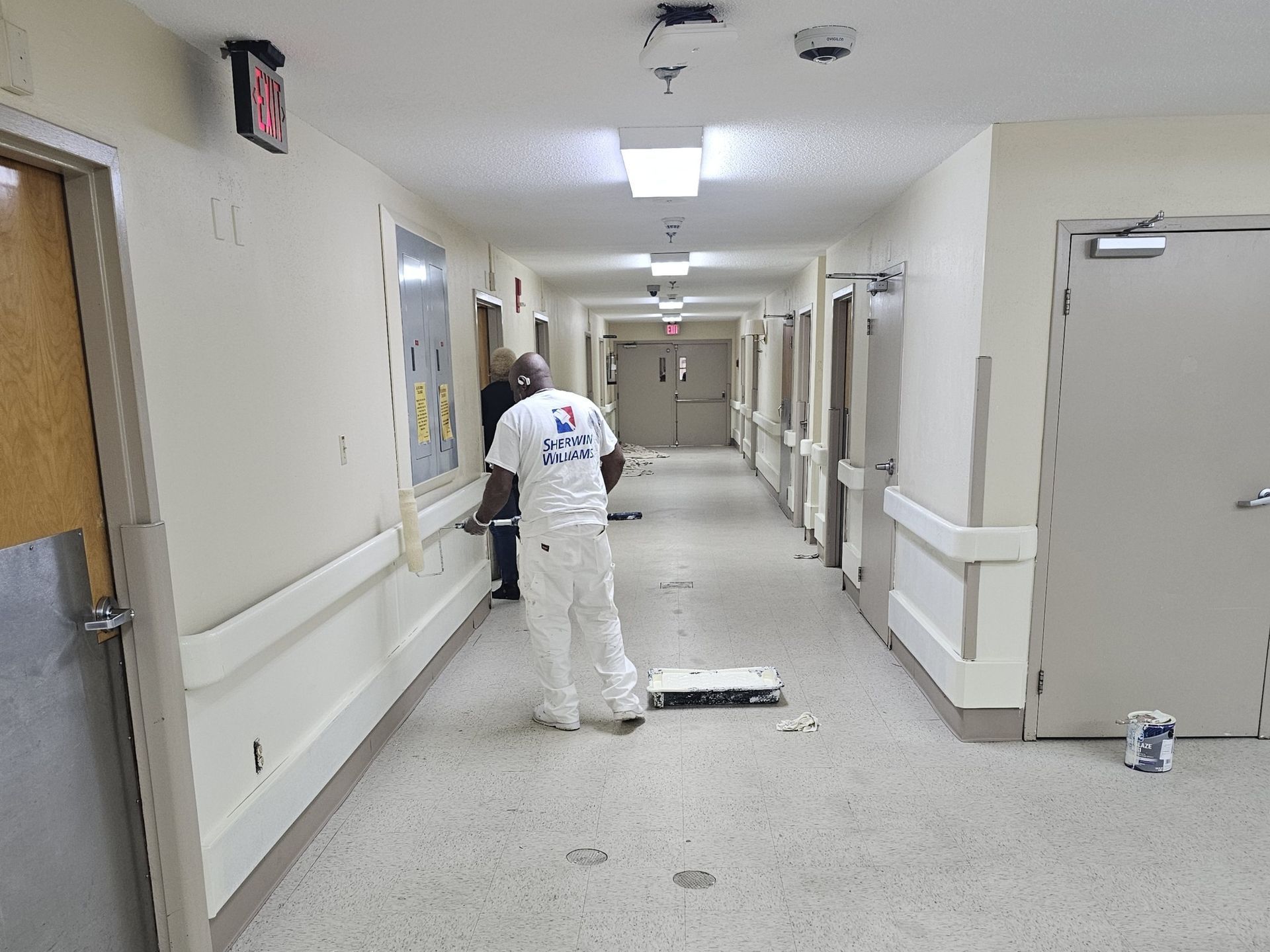 Two workers painting a long hallway with white walls, doors, and handrails.
