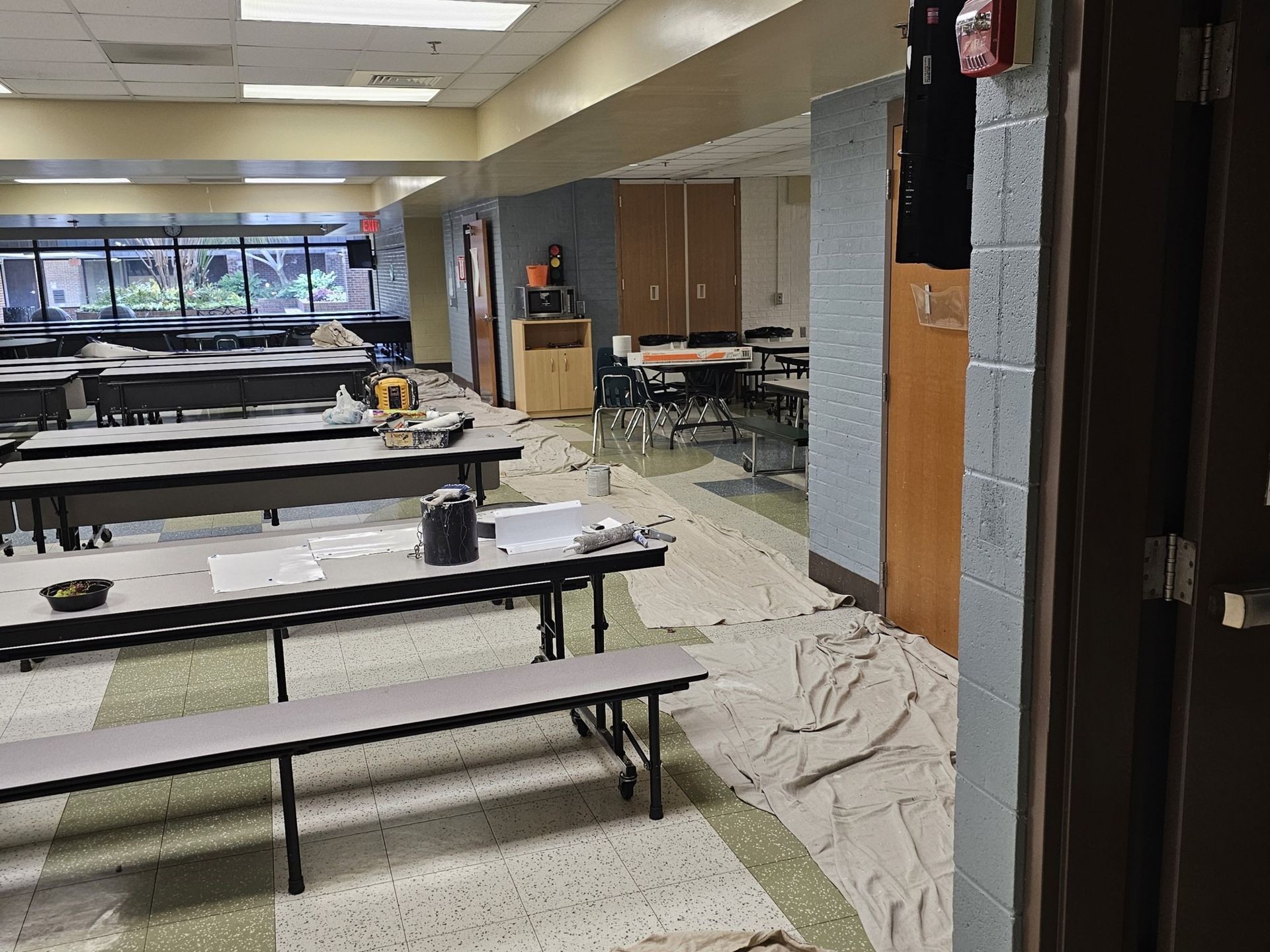 School cafeteria during renovation, tables covered, paint supplies, white and gray walls.